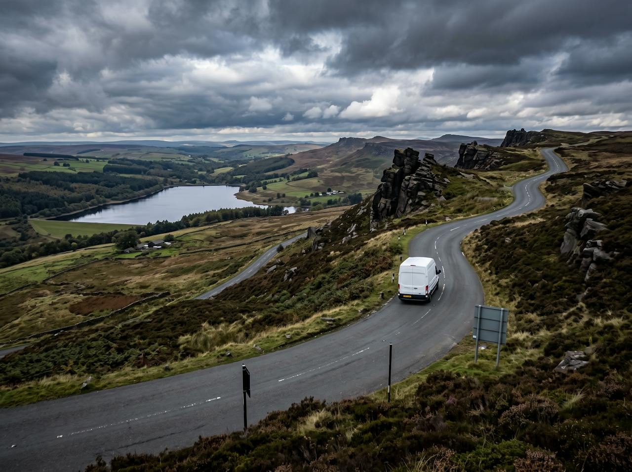 White van on a winding moorland road past gritstone outcrops and a reservoir near Leek